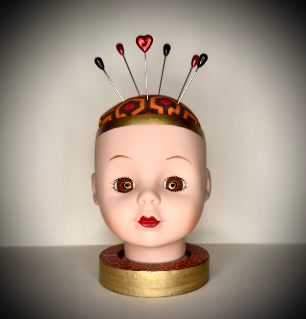 Doll head pin cushion bust with large brown eyes, red lips, with a Hicks Hexagon patterns cushion and base.